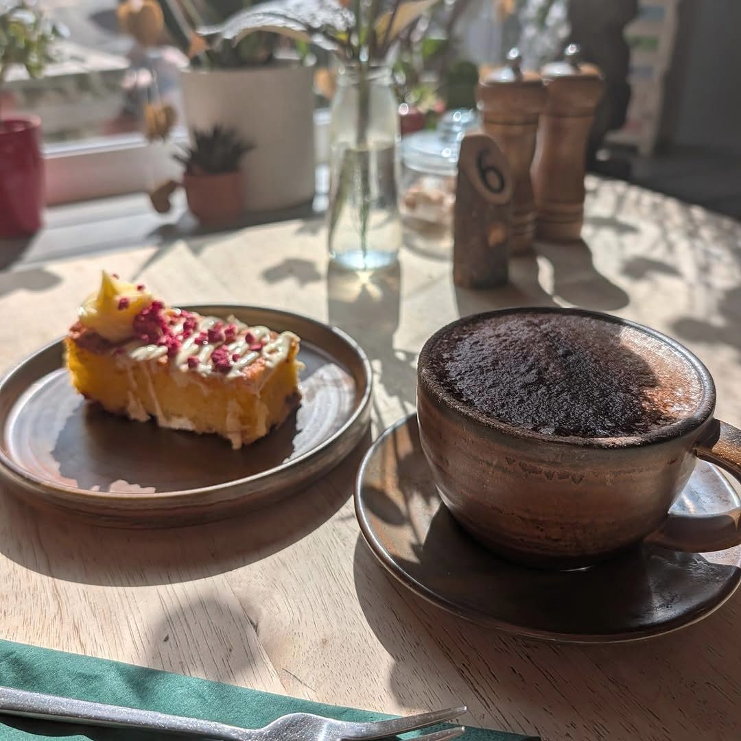 A mocha in a brown stoneware cup beside a slice of homemade cake, on a wooden table by the window