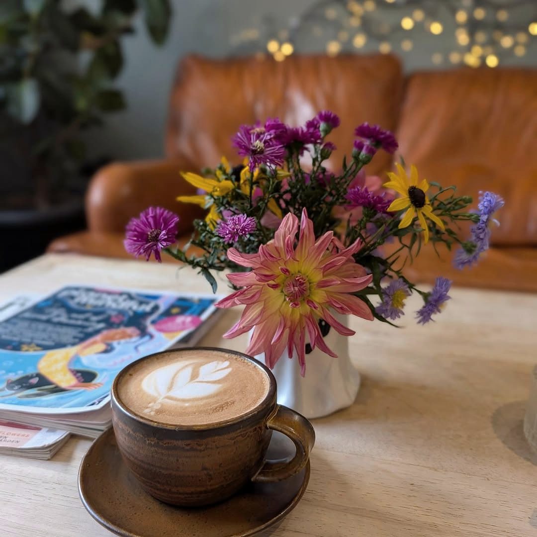 A flat white with latte art in front of a jar of garden flowers on a wooden table