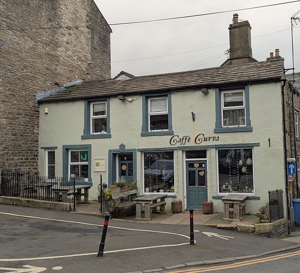 Caffè Curva on a corner in Hawes: a pale rendered building with grey-blue window frames, the café's name above the windows, benches outside and the road curving past
