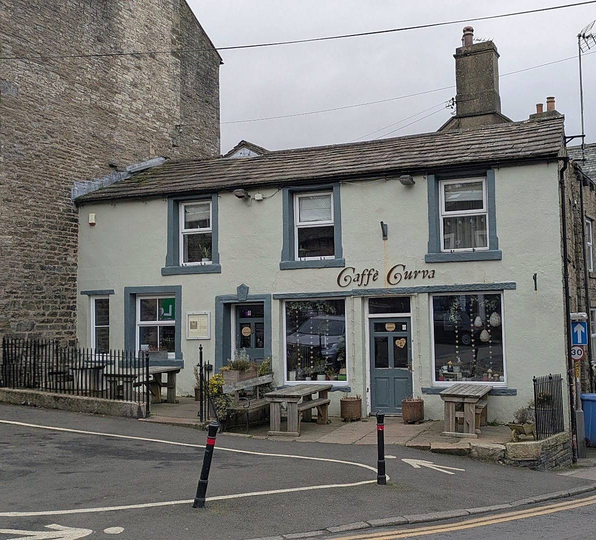 Caffè Curva on a corner in Hawes: a pale rendered building with grey-blue window frames, the café's name above the windows, benches outside and the road curving past