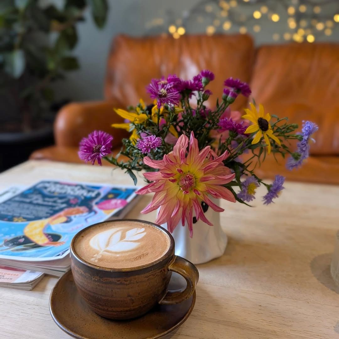 A flat white with latte art in front of a jar of garden flowers on a wooden table